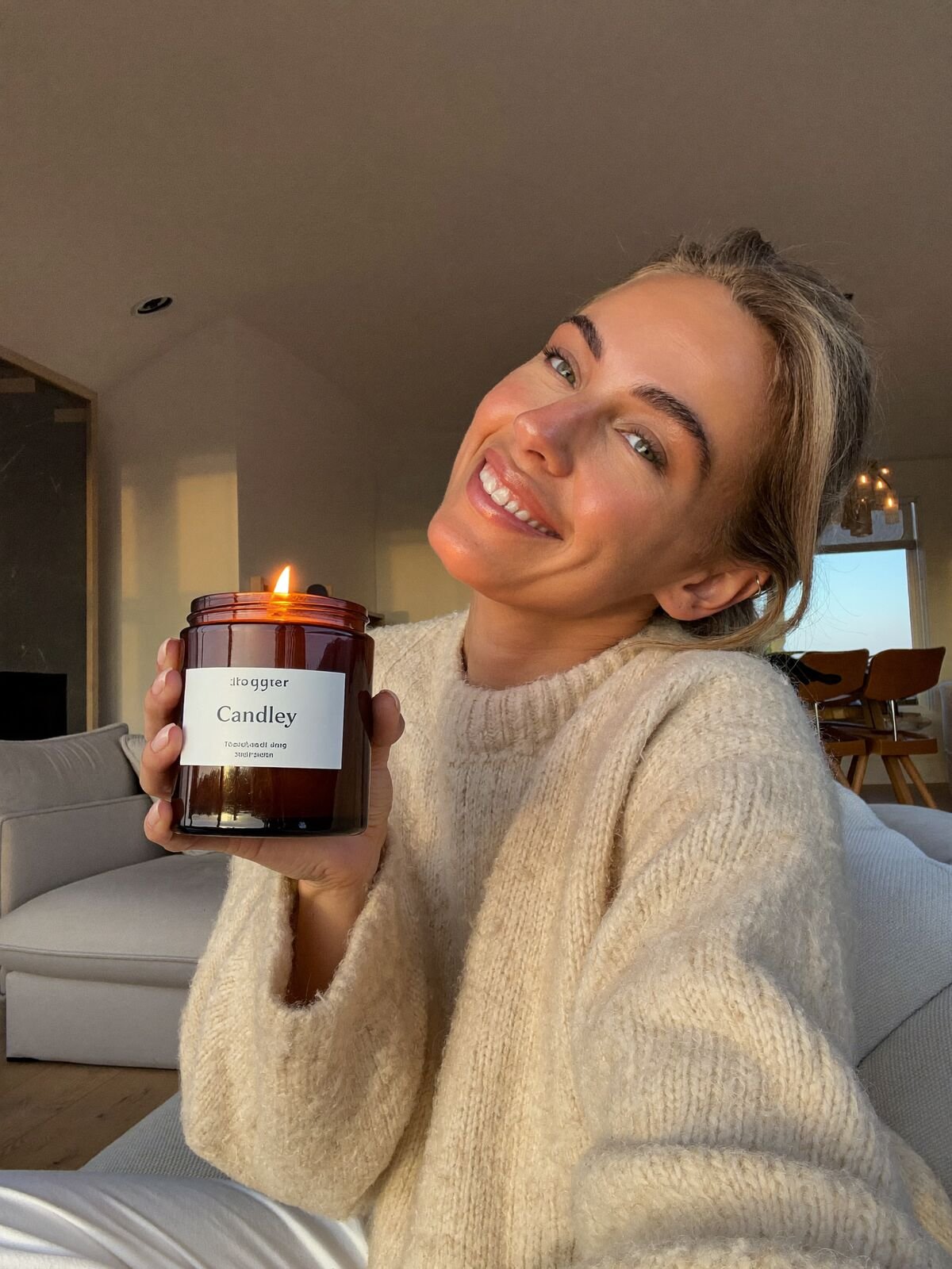 Smiling woman in a knit sweater holding a lit candle in a cozy living room