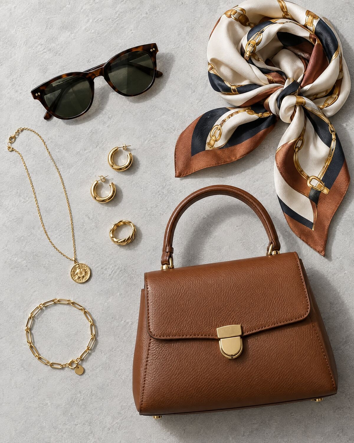 Overhead flat lay of gold jewelry, a leather handbag, sunglasses and a silk scarf