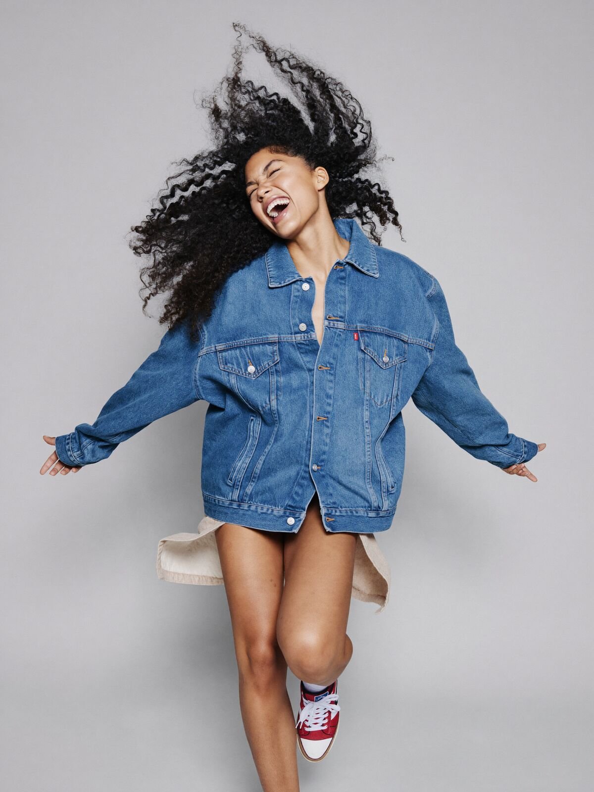 Young woman laughing mid-spin in a denim jacket and red sneakers