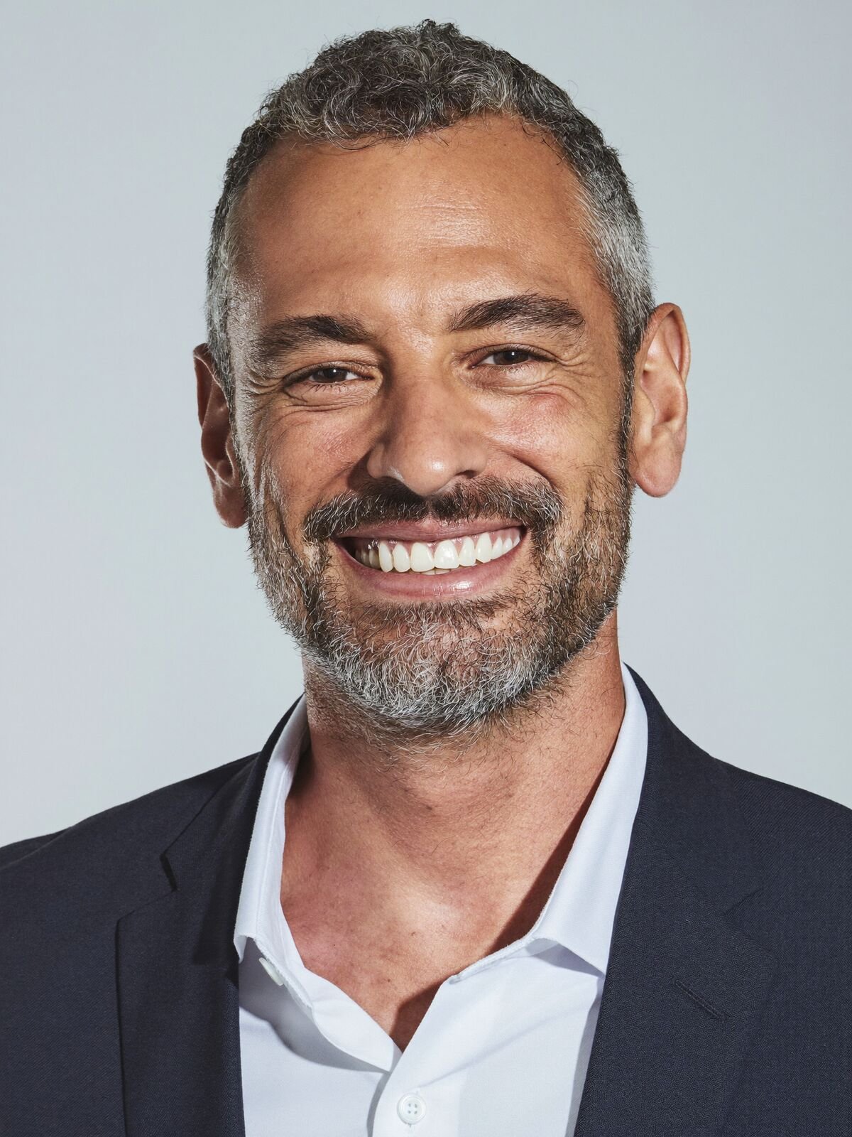 Smiling man with a salt-and-pepper beard in a navy jacket on a light grey backdrop